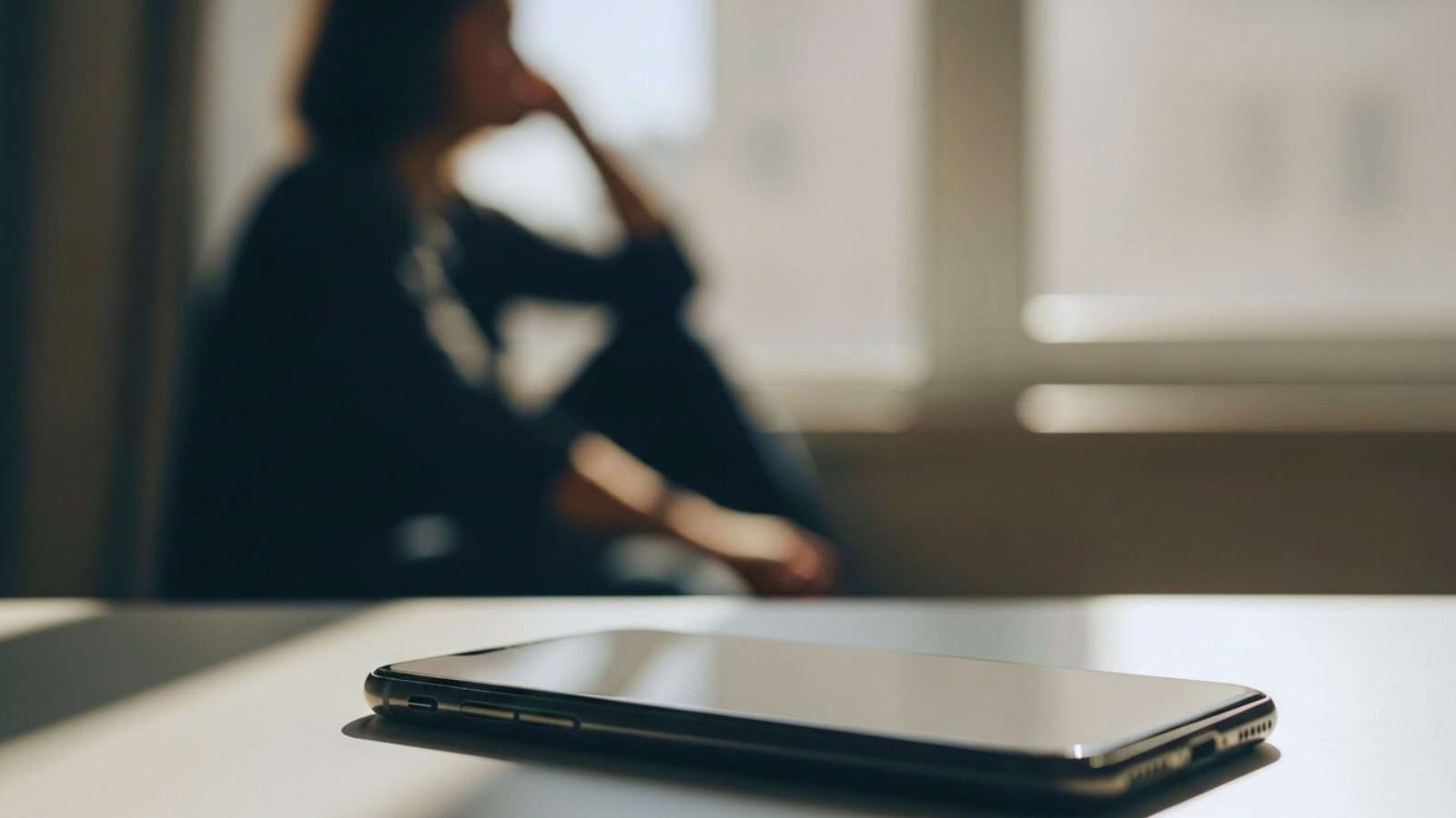 Teléfono móvil sobre una mesa en primer plano, con una figura humana desenfocada al fondo, en un ambiente de reflexión y pausa